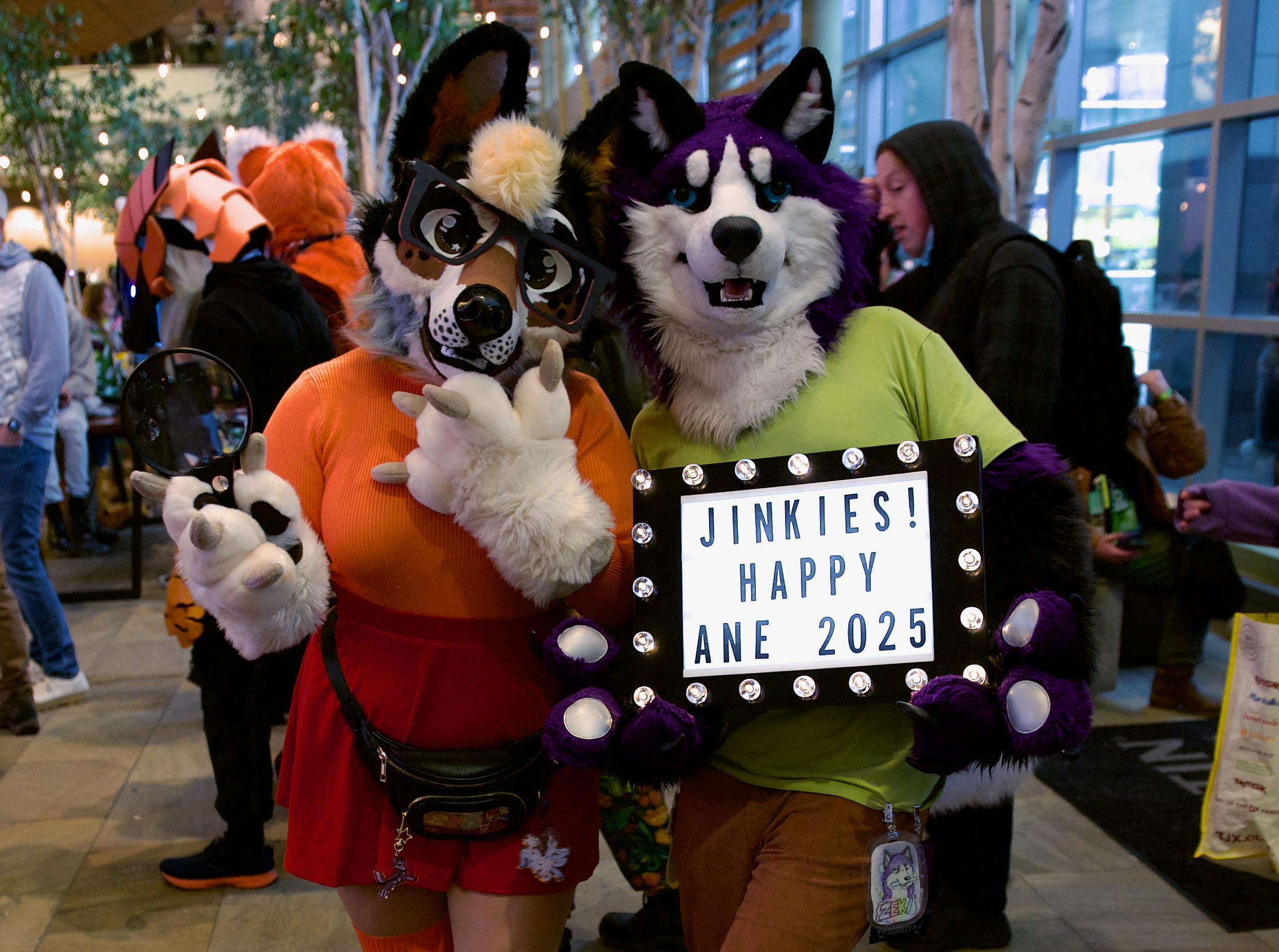 Two fursuiters cosplay as Shaggy and Velma from the Scooby Doo Franchise. Shaggy holds up a sign saying "Jinkies! Happy ANE 2025".